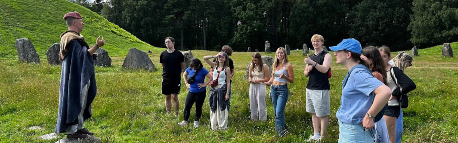 Eleven students, listening to a tour guide, while standing in a grassy field filled with large stones.